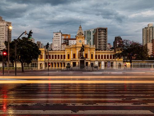 Foto noturna de longa exposição. O edifício da estação, iluminado em amarelo, está ao fundo. Em primeiro plano, a rua molhada e a faixa de pedestres. Rastros de luz horizontais, de faróis de carros, cruzam a imagem em tons de branco e vermelho, mostrando o movimento do trânsito.