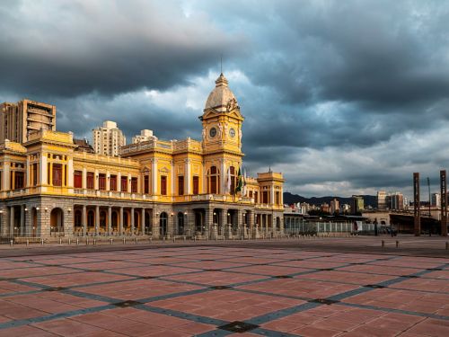 Vista ampla da praça com seu piso de ladrilhos vermelhos e detalhes em cinza. À esquerda, o imponente edifício histórico da estação, de cor amarela, sob um céu dramático, com nuvens cinzentas e carregadas. Ao fundo, à direita, veem-se outras estruturas urbanas e serras no horizonte.
