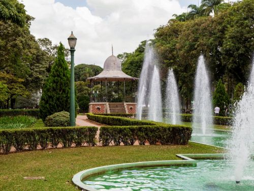 Coreto histórico de ferro com teto cônico e cúpula metálica ao centro da praça, acessível por escadaria de tijolos decorados. Em frente, fonte retangular com jatos d’água altos sobre lago verde. Caminhos de terra, postes verdes clássicos e jardins podados completam o cenário arborizado.