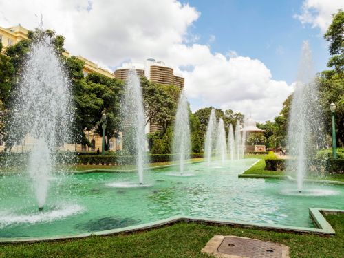 Fonte retangular de água verde com fileira de jatos altos alinhados. À esquerda, prédio histórico amarelo com janelas arqueadas; ao fundo, edifício moderno arredondado contrasta com o coreto histórico ao centro. Céu parcialmente nublado, árvores e jardins enquadram a cena.