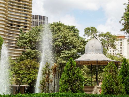 Dois jatos de fonte vertical em primeiro plano à esquerda; área ajardinada com arbustos e canteiros baixos ao centro. À direita, quiosque/gazebo metálico com cúpula e plataforma elevada com degraus. Árvores de copa densa e prédios altos parcialmente visíveis ao fundo.