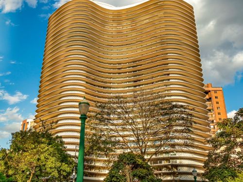 Edifício modernista de linhas onduladas e varandas contínuas brancas contrasta com o céu azul e nuvens claras. Em primeiro plano, poste verde clássico de luminária, banco de ferro e árvores que projetam sombras no caminho de pedras. Jardins floridos e arbustos completam o espaço urbano arborizado.