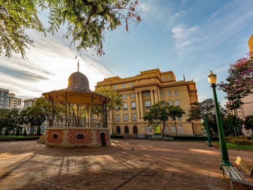 Coreto circular de ferro fundido com colunas e teto cônico de telhas cinzas, cercado por bancos e árvores. Piso em pedras vermelhas. Ao fundo, prédio histórico amarelo com colunas brancas e janelas altas. Luminárias verdes completam o cenário urbano.