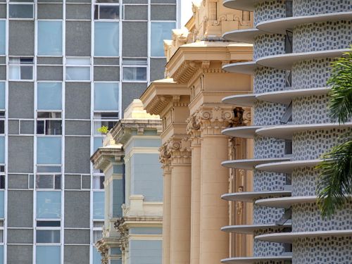 Contraste arquitetônico: à esquerda, fachada antiga bege com colunas e ornamentos; ao centro, edifício moderno cinza com janelas retangulares; à direita, prédio curvo azul com varandas brancas horizontais. Paisagem urbana de estilos sobrepostos.