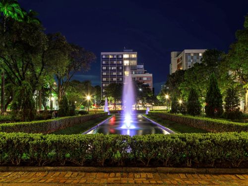 Fonte retangular iluminada com jatos verticais coloridos em tons de roxo e azul. Em volta, jardins podados e árvores escuras. Ao fundo, prédios residenciais com luzes acesas. Reflexos vibrantes na água marcam o clima noturno.