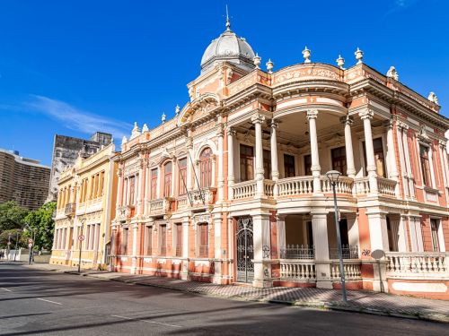 Edifício histórico rosa com fachada ornamentada em branco. No topo, cúpula metálica arredondada. Varanda central com colunas e balaústres brancos, janelas de madeira fechadas. Em frente, rua asfaltada; atrás, prédios modernos contrastam o estilo.
