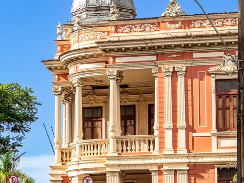 Fachada detalhada de prédio histórico rosa. Varanda semicircular com colunas brancas e balaustrada. Cúpula metálica prateada no topo, ornamentos em relevo coroam a estrutura. Estilo clássico preservado, com cores contrastantes e aspecto imponente.