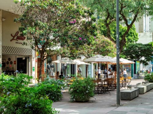 Calçada larga arborizada com mesas e cadeiras de madeira sob guarda-sóis brancos. Fachada de sorveteria à esquerda. Vaso com arbustos verdes e árvores floridas distribuem-se pelo espaço. Prédios altos ao fundo.