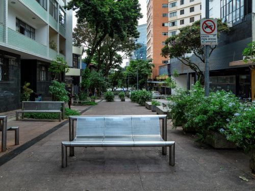 Calçadão pavimentado com bancos metálicos prateados no centro e vasos de concreto com arbustos verdes. Prédios residenciais e comerciais cercam a praça. Sinalização de trânsito indica acesso restrito a garagens.