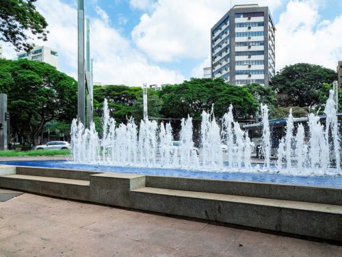 Fonte com jatos de água verticais em tanque retangular azul. Bancos de concreto envolvem a estrutura. Ao fundo, árvores verdes e prédios altos. Parte do céu aparece nublado.