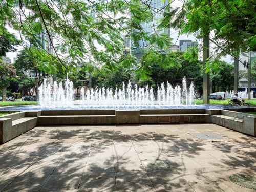 Fonte retangular com jatos de água brancos ao centro. Em volta, bancos de concreto e calçamento de pedras. Árvores grandes projetam sombra no chão. Carros circulam no fundo da praça.