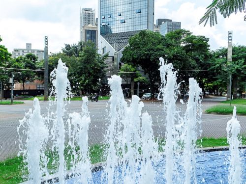 Fonte retangular de água azul em primeiro plano, jatos verticais brancos no centro. Ao fundo, prédio alto de vidro espelhado e árvores verdes. Céu parcialmente nublado.