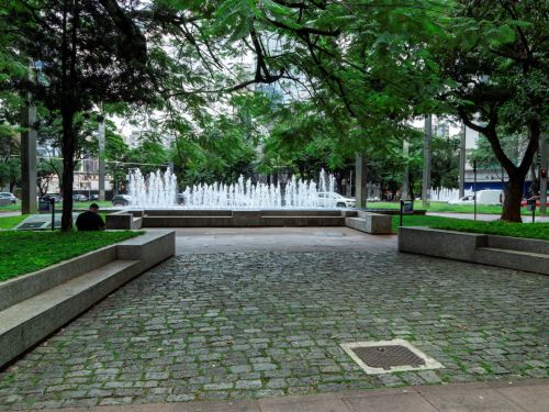 Fonte com jatos de água brancos cercada por bancos de concreto. Calçamento de pedras no chão e árvores verdes nas laterais. Pessoas caminham no canto esquerdo. Carros e prédios aparecem ao fundo.