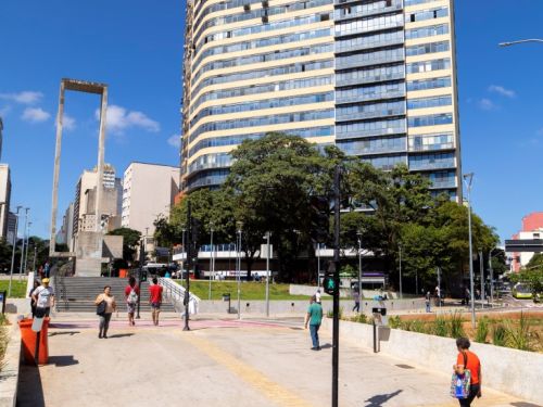 Em primeiro plano, uma faixa de pedestres com largas listras vermelhas e brancas. Pessoas caminham pela praça. Ao fundo, um grande edifício residencial com fachada de cor creme e janelas espelhadas. À esquerda, destaca-se o monumento de concreto da praça, sob um céu azul intenso.