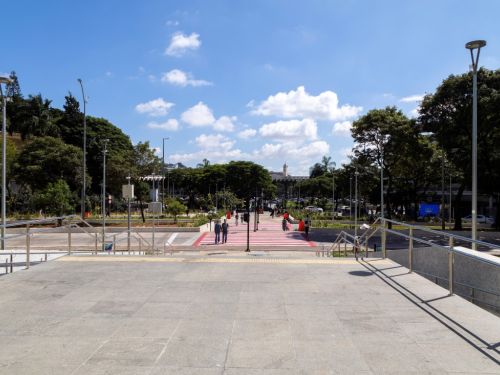 Vista do topo de uma escadaria de granito, olhando para a praça. Em frente, uma faixa de travessia de pedestres rosa e branca. A praça é arborizada, com canteiros de grama. Ao fundo, no centro, o prédio da prefeitura com sua torre do relógio se destaca entre as árvores. O céu está azul.