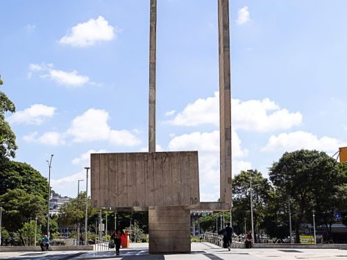 O monumento de concreto da praça é visto de frente, a partir de um pátio de cimento claro. A escultura tem uma grande base retangular e duas colunas altas que se conectam no topo, formando um portal vazado. Pessoas caminham ao redor. O céu é azul com nuvens brancas e esparsas.