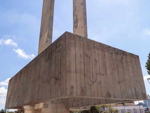Fotografia em de baixo pra cima do grande monumento de concreto da praça. A base retangular e robusta se projeta sobre pilares menores. As duas colunas altas sobem em direção ao céu. O sol brilha intensamente no canto superior direito, criando um brilho forte contra o céu azul claro.