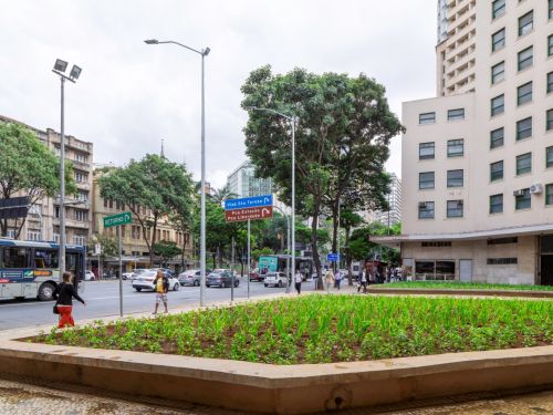 Vista de praça urbana com canteiro retangular de plantas verdes no centro. À esquerda, ônibus azul e branco, árvores e prédios antigos. À direita, edifício alto com fachada clara e janelas verdes. Pessoas caminham pela calçada.