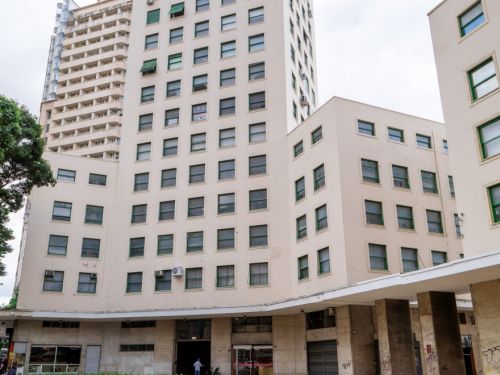 Edifício alto e claro, de vários andares, com janelas de moldura verde. Na frente, canteiro de plantas recém-cultivadas. O prédio possui base com colunas largas e espaço de passagem coberto. Céu nublado ao fundo.