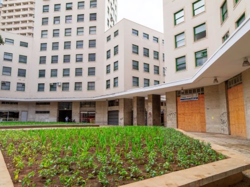 Prédio claro com janelas verdes formando conjunto arquitetônico em “U”. No centro, canteiro de plantas verdes e caminhos de pedra. Parte do térreo tem portas de madeira vedadas e paredes com pichações.