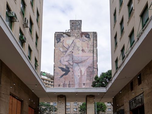 Corredor externo entre prédios claros com janelas verdes. No centro, cobertura sustentada por colunas. Ao fundo, mural grande pintado em prédio vizinho, representando duas figuras humanas. Piso de pedras e paredes laterais com portas fechadas.
