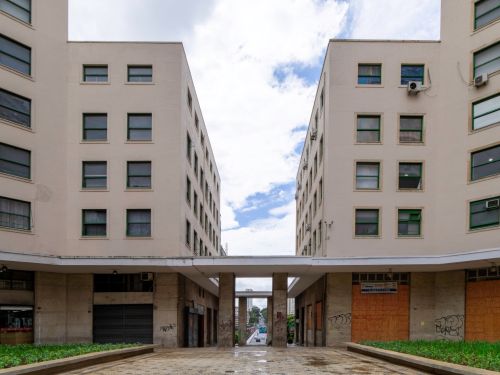 Vista simétrica de dois blocos de prédio claros, paralelos, com janelas verdes. No centro, passarela coberta por colunas. À frente, chão de pedras e canteiros laterais. Portas no térreo estão fechadas e há pichações nas paredes.
