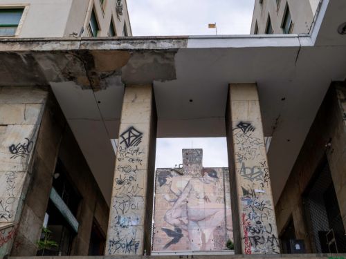 Escadaria de pedra em primeiro plano, com paredes e colunas cobertas por pichações. Acima, cobertura branca sustentada por pilares. Ao fundo, grande mural em prédio vizinho, mostrando duas figuras humanas em tons claros.