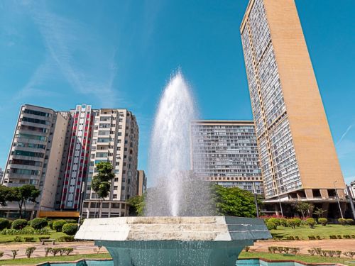 Um jardim com flores vermelhas vibrantes preenche a parte inferior da imagem. Logo acima, um edifício alto em estilo art déco, de cor marrom-alaranjada, se destaca. Ao seu lado, outro prédio de fachada clara. O céu ao fundo é de um azul intenso, com uma pequena nuvem passando.
