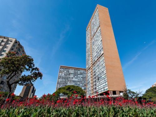 Um caminho largo e pavimentado com pedras claras apresenta desenhos em espiral feitos com pedras mais escuras, criando um efeito visual que conduz o olhar. O caminho é ladeado por gramados verdes e arbustos baixos e bem cuidados. Ao fundo, altos edifícios da cidade sob um céu azul e limpo.