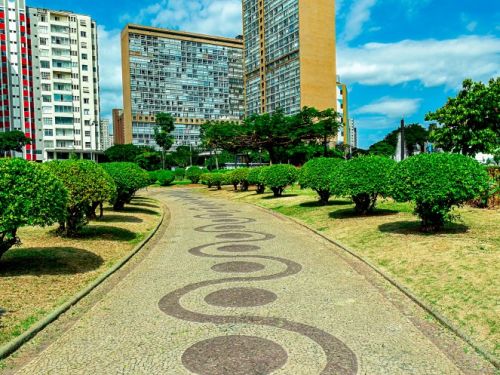 A partir de um canteiro gramado, a cena mostra arbustos verdes podados em formato de bola. Uma árvore projeta sombras sobre a grama. Ao fundo, vários edifícios altos de diferentes estilos e cores, e ao centro, a fonte da praça com seus jatos de água ativos à distância.