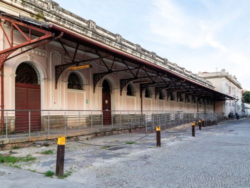 Fachada de um prédio antigo em tom rosa claro, com portas e janelas em arco, algumas gradeadas. A entrada tem uma pequena rampa com corrimão metálico, e o passeio é de pedra clara. Há sombra projetada na calçada, sugerindo árvores próximas.
