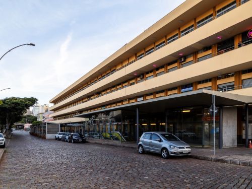 Trecho de rua pavimentada com paralelepípedos, onde alguns carros estão estacionados ao longo da calçada. À esquerda há um prédio amarelo claro, de vários andares, com marquise sobre a calçada e portas comerciais no térreo.