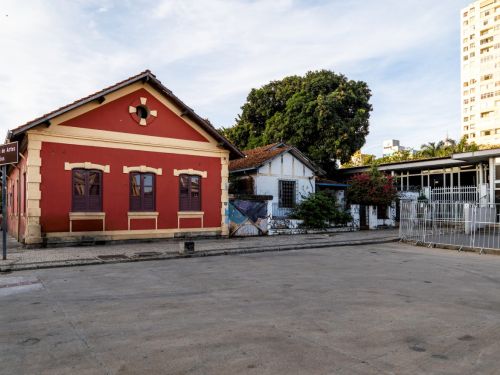 Casa térrea pintada de vermelho, com janelas de madeira e telhado de duas águas. Ao lado, uma construção branca de aparência mais simples. Árvores aparecem no fundo, atrás das edificações.