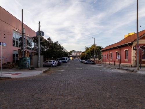 Rua estreita calçada com paralelepípedos. À direita, uma casa vermelha com portas e janelas de madeira. À esquerda, um prédio de fachada rosa clara com grades. A via segue levemente em curva, e há postes de iluminação.