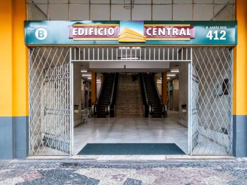 Entrada de uma galeria comercial, identificada por uma placa com o nome “Edifício Central”. Escadas rolantes descem logo na entrada, e o teto é baixo, com iluminação artificial. Há pessoas circulando ao fundo.