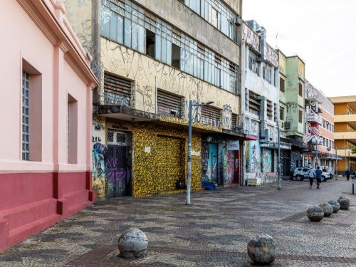 Fachadas de prédios baixos e antigos, desgastados pelo tempo. Algumas superfícies estão grafitadas. O passeio tem pequenas esferas de concreto servindo como barreira para veículos, e há postes e fios elétricos atravessando o cenário.