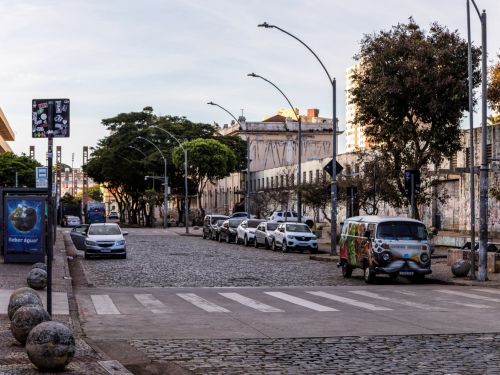 Rua larga e plana, calçada com paralelepípedos, onde alguns carros estão estacionados paralelamente. No fundo, vê-se prédios antigos de médio porte, com varandas e janelas alinhadas.