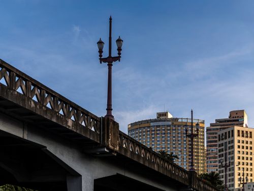 Viaduto de concreto com muretas e postes antigos de iluminação. Ao fundo, surgem prédios altos de diferentes estilos arquitetônicos, alguns mais modernos. 
