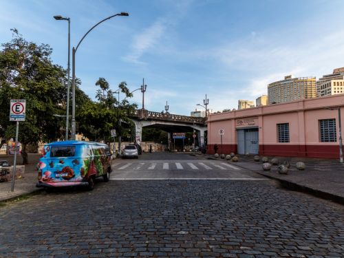 Rua de paralelepípedos com faixa de pedestres pintada de branco. Uma van colorida está estacionada no lado direito, próxima a uma calçada larga. Ao fundo, vê-se um viaduto elevada cruzando a paisagem.