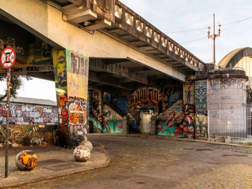 Imagem de um viaduto com pilares e paredes cobertos por grafites coloridos. Há escadarias sob o viaduto e bolas de concreto pintadas no chão. Um poste com luminária antiga está posicionado acima da estrutura.