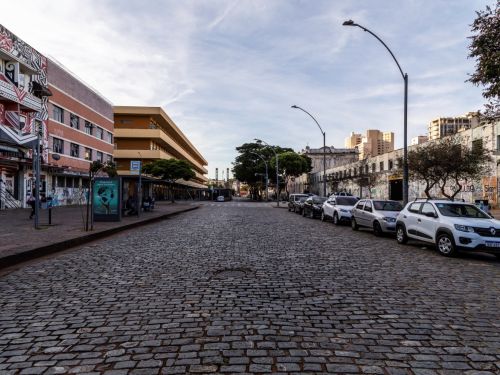 Rua calçada com paralelepípedos, com carros estacionados em uma das laterais. Do lado esquerdo, há prédios com fachadas coloridas e ponto de ônibus. O lado direito possui um muro grafitado e árvores pequenas.
