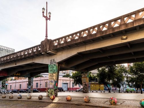 Vista ampla do vão sob o Viaduto Santa Tereza. Os robustos pilares de concreto que sustentam a estrutura estão cobertos por grafites. Um poste de iluminação de cor vinho se ergue em frente a um dos pilares. No chão, uma calçada com esferas de concreto pintadas artisticamente.