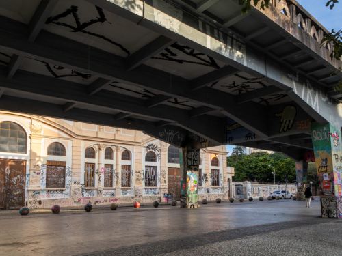Vista sob o viaduto mostrando o piso de concreto e pilares grafitados. Ao fundo, há um prédio antigo com fachada clara e janelas arqueadas, coberto por pichações e grafites.