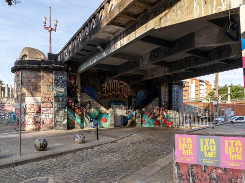 Imagem mostra a base de um viaduto grafitado com duas escadas laterais que levam a uma porta central. Há um poste com luminária antiga e bolas de concreto pintadas espalhadas na calçada.