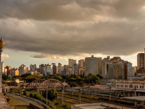 Vista do horizonte de Belo Horizonte a partir de um ponto elevado. Em primeiro plano, trilhos de trem e o Viaduto de Santa Tereza, com seus arcos de metal. Ao fundo, diversos prédios do centro da cidade sob um céu com nuvens densas e escuras, iluminadas por uma luz dourada de fim de tarde.