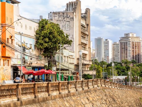 Vista a partir do muro de arrimo da rua. Um gradil de cimento ornamentado está em primeiro plano. Logo atrás, há um bar com mesas e guarda-sóis vermelhos. Ao fundo, edifícios de diferentes estilos, incluindo um prédio antigo com pichações, e o horizonte da cidade com prédios modernos.