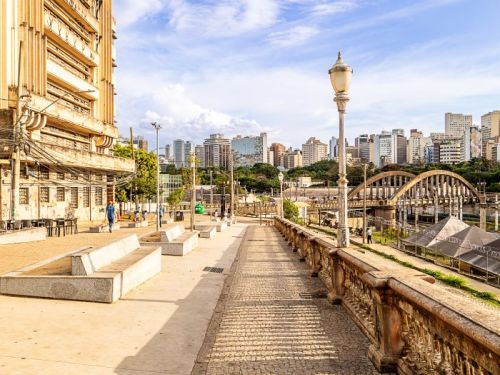 Perspectiva de uma calçada longa, parte em cimento e parte em pedras portuguesas. À direita, um gradil de cimento ornamentado e um poste de iluminação republicano. À esquerda, um grande edifício antigo e bancos de concreto. Ao fundo, o Viaduto de Santa Tereza e o centro da cidade.