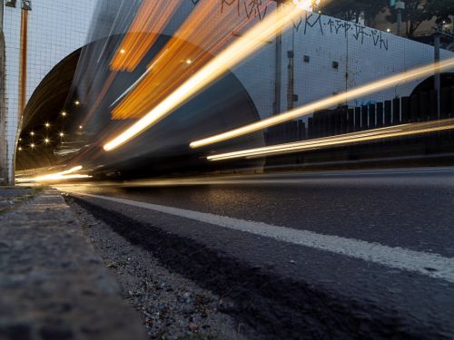 Foto de longa exposição em ângulo muito baixo, a partir da guia da calçada. A entrada do túnel em arco está à esquerda. Feixes de luz grossos, em tons de laranja e branco, parecem explodir de dentro do túnel, projetando-se em diagonal para fora e para o alto, cruzando a imagem inteira.