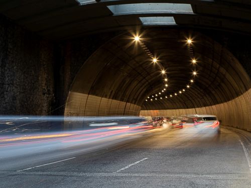 Visão de dentro do túnel, que tem formato de arco com paredes de concreto. Luzes no teto formam um caminho curvo. Carros em movimento se transformam em rastros de luz borrados, predominantemente em tons de vermelho e branco, criando uma forte sensação de velocidade ao longo da via.