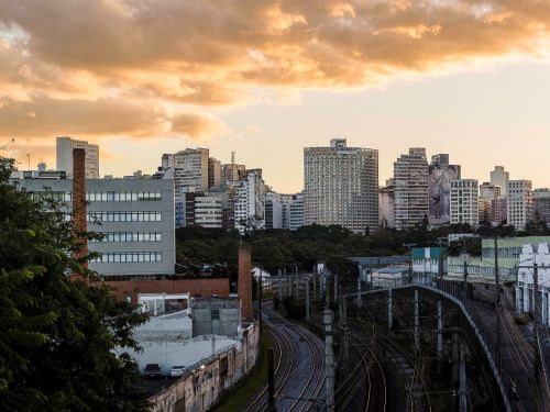 Paisagem urbana vista do alto. Em primeiro plano, múltiplos trilhos de trem que fazem curvas suaves. À esquerda, um muro e uma chaminé de tijolos. Ao fundo, o horizonte é preenchido por diversos prédios de diferentes alturas e estilos. O céu está coberto por nuvens de cor alaranjada.